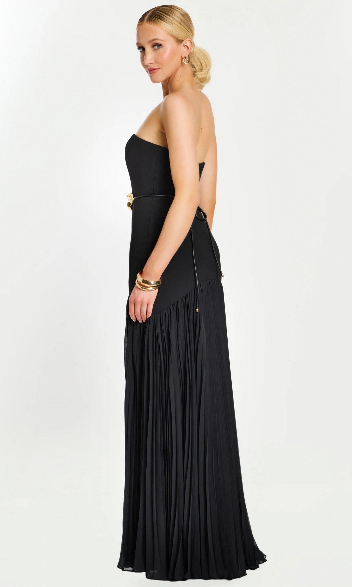 A woman in a strapless black Jovani 47000 floor-length formal dress featuring a pleated skirt and gold floral waist detail, accessorized with gold jewelry, stands against a plain light background.